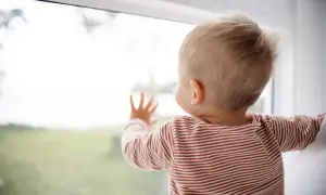 Niemowlę w paski patrzy przez okno, dotykając szyby dłonią.