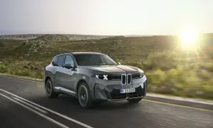 El BMW iX5, un SUV eléctrico gris, avanza por una carretera rural al atardecer, con colinas verdes y el sol brillando.
