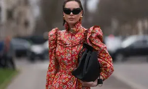 Kobieta w sukience w kwiaty, z bufiastymi rękawami i falbankami, prezentuje styl romantyczny. Czarna torebka i okulary dodają jej charakteru.