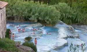 Persone si rilassano nelle cascate naturali dei aspio terme, con vegetazione lussureggiante e un antico edificio in pietra.