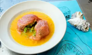 En skål med fisk och grönsaker i en gul vätska, kanske en läcker leche de tigre. Två ostronskal ligger på ett blått underlägg med en hajritning.