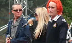 Ich troje wokalistki na scenie, z mikrofonami w dłoniach. Jeden z nich ma ogniste, pomarańczowe włosy.