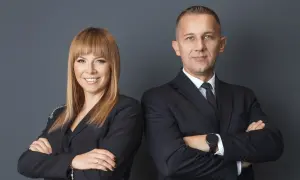 Dwie osoby w czarnych garniturach, kobieta i mężczyzna, stoją obok siebie ze skrzyżowanymi rękami.