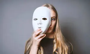 Una ragazza con lunghi capelli biondi tiene una maschera bianca davanti al viso. Forse un istrionico può amare, ma nasconde il suo vero volto.
