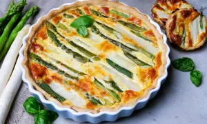 Herzhafte Spargel Quiche mit frischem Spargel und Kräutern, goldbraun gebacken. Daneben liegen roher Spargel und kleine Quiches.