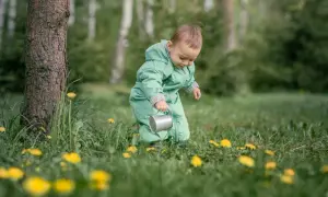 Małe dziecko w zielonym kombinezonie bawi się konewką wśród traw i żółtych kwiatów.