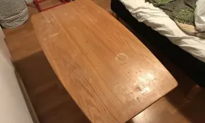 Ett slitet soffbord i trä med fläckar, redo att måla om. Bredvid syns en säng med vitt och grönt överkast.