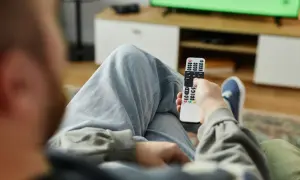 Mężczyzna z pilotem w ręku, zastanawia się, kto nie musi płacić abonamentu RTV, oglądając zielony ekran telewizora.