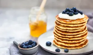 Pile de pancakes moelleux, garnis de crème et de myrtilles, avec un pot de miel en arrière-plan. Une recette pancake parfaite pour le petit-déjeuner.