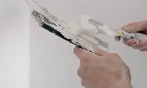 Händer som spacklar en vägg. Spacklet är vitt och klibbigt, och verktygen är täckta av det.