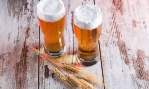 Dwa kufle złocistego piwa, przypominającego red ale, z obfitą pianką, stoją na drewnianym blacie obok kłosów zboża.