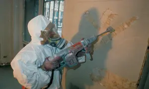 Travailleur en combinaison intégrale et masque à gaz, utilisant un marteau-piqueur pour retirer un revêtement mural. Que faire face à l'amiante au travail.