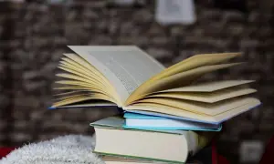 Otwarta książka leży na stosie innych książek, na tle ceglanej ściany.