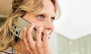 Kobieta z jasnymi włosami i niebieskimi oczami rozmawia przez telefon. Jej dłoń z pierścionkami ozdabia urządzenie.