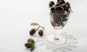 Un bicchiere pieno di olive nere, con rametti e foglie, accanto a olive fresche su un pizzo. Le olive delle Marche, un sapore autentico.