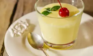 Puszysty deser kogel-mogel w szklance, udekorowany wiśnią i miętą, na talerzyku z łyżeczką.