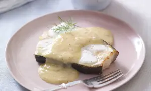 En bit fisk täckt med krämig beurre blanc-sås, garnerad med dill, serveras på en ljusrosa tallrik med en gaffel.