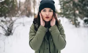 Czarna czapka świetnie komponuje się z zieloną kurtką. Dziewczyna w zimowym lesie, z dłońmi przy twarzy.