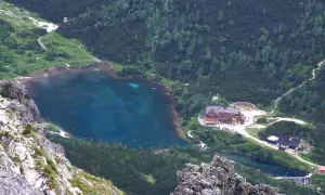 Widok na zielony staw kieżmarski otoczony lasem i górami. W pobliżu schronisko i ścieżki.