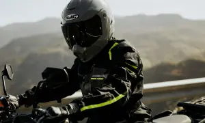 Motocyklista w szarym kasku HJC i czarnej kurtce z neonowymi detalami jedzie motocyklem po górskiej drodze.