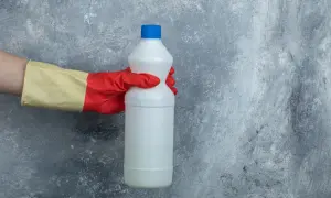 Mano con guanto rosso e giallo tiene una bottiglia di candeggina, pronta per la lavatrice.