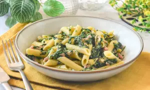 Un plato de pasta penne con espinacas y trocitos de jamón, servido en un cuenco sobre una servilleta amarilla.