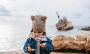Małe dziecko w czapce z pomponami bawi się na plaży na Cyprze, w tle wrak statku.