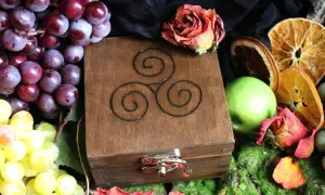 Drewniana szkatułka z symbolem triskelion, otoczona winogronami, suszonymi pomarańczami, jabłkiem i różą.