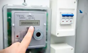 Palec wskazuje licznik energii elektrycznej, pokazujący 717.0 kilowatogodzina. Obok widoczny jest wyłącznik nadprądowy.