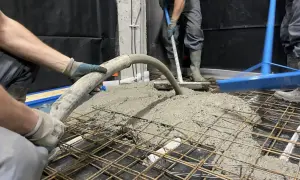 Pracownicy wykonują pompowanie betonu na zbrojenie. Beton płynie z węża, a drugi pracownik rozprowadza go łopatą.
