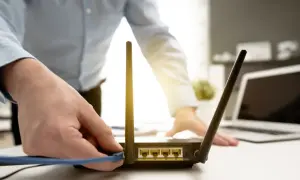 Dłoń podłącza kabel do routera, co może być pierwszym krokiem, jak zresetować router. W tle widać laptopa.