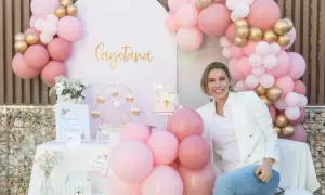 Una mujer sonríe junto a una mesa decorada para una comunión casera, con globos rosas y dorados, un pastel y un cartel de bienvenida.