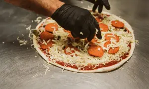 Händer i svarta handskar lägger ost på en pizza med pepperoni och oliver. En klassisk kombination för härliga pizza smaker.