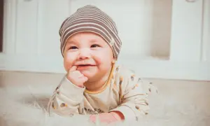 Uśmiechnięte dziecko w czapce leży na białym futerku, z palcem w ustach.