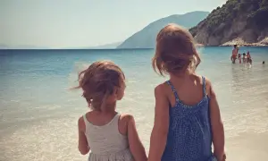 Dwie dziewczynki trzymają się za ręce na plaży w Albanii, patrząc na morze i góry. W tle w wodzie bawią się inni ludzie.