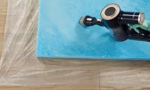 A gloved hand uses a vacuum cleaner with a brush attachment to clean a blue foam mattress topper laid on plastic sheeting.