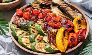 Una colorida parrillada de verduras con calabacín, berenjena, tomates cherry, pimientos y cebolla, servida en una bandeja de madera.
