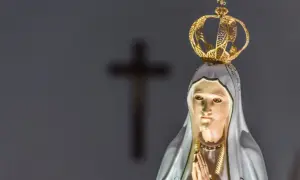 Figurka Matki Bożej z Fatimy, w koronie, z modlącymi się dłońmi. W tle niewyraźny krzyż, symbol objawienia fatimskiego.