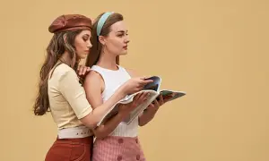 Dwie kobiety w stylizacjach inspirowanych **modą lat 60.** przeglądają magazyn. Jedna w skórzanym beretku, druga w opasce na włosach.