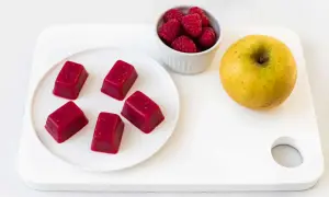 Cinq carrés de pâte de fruit rouge, des framboises et une pomme jaune sur une planche blanche. Une idée de conservation des fruits.