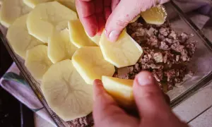 Dłonie układają plastry ziemniaków na mielonym mięsie, tworząc prostą zapiekankę ziemniaczaną.