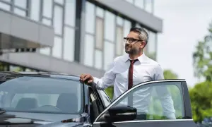 Biznesmen w białej koszuli i krawacie opiera się o drzwi czarnego samochodu. Czeka na niego kolejna kilometrówka.