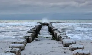 Drewniane pale na plaży, fale rozbijające się o brzeg, pochmurne niebo.