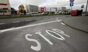 Na asfalcie widoczna jest biała linia warunkowego zatrzymania "STOP". W tle budynki i ruch uliczny.