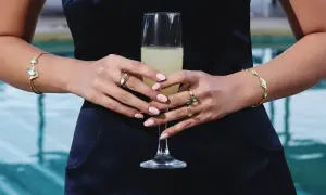 Manos con uñas rosadas y varios anillos dorados, sosteniendo una copa de champán. Brazaletes dorados adornan ambas muñecas.