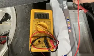 Multímetro amarillo muestra 10.90V al comprobar bateria coche. Se ve la batería, un neumático y cables.