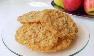 Tre gyllene glutenfria havreflarn ligger staplade på en glasfat. I bakgrunden syns äpplen och päron.