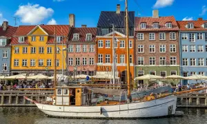 Ein weißes Segelschiff liegt im Hafen vor bunten Häusern. Menschen sitzen in Cafés am Wasser. Ein perfekter Tag im kopenhagen sommer.
