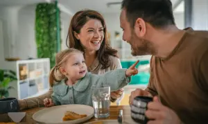 Szczęśliwa rodzina przy stole, jedząca pizzę. Dziecko wskazuje palcem, jakby opowiadało o przyszłym domu, marząc o rodzinnym kredycie mieszkaniowym.