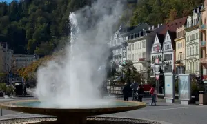 Der sprühende geysir karlsbad mit dampfender Fontäne vor bunten Häusern und grünen Bäumen.